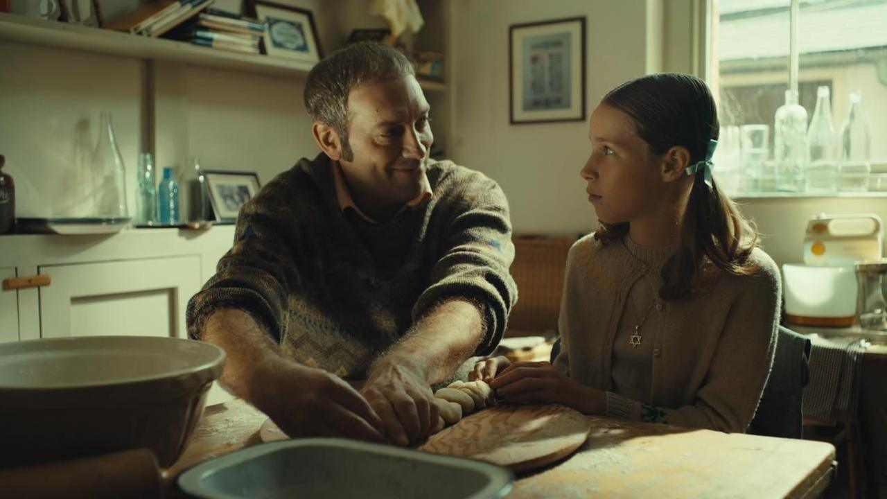 A still from a film of a man smiling as he looks across to a young girl while they sit at a table in an old-fashioned kitchen.