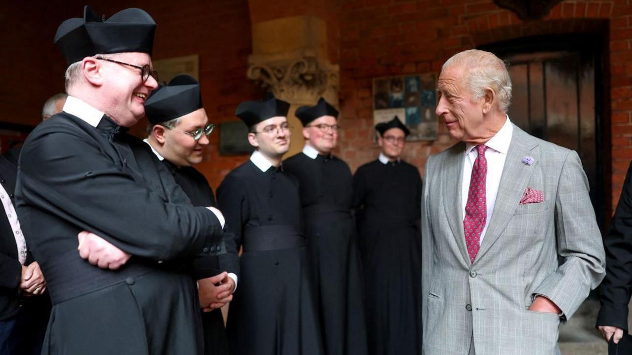 The King, in grey suit and pink tie chats to a Father in a black coat, with white collar and black pointed hat. Four male colleagues in the same outfits look on. They are inside a building which has exposed red brick walls and ornate stone carvings on pillars.