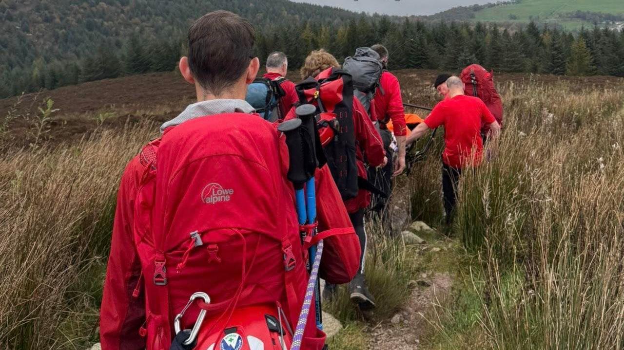 A group of mountain rescuers, viewed from behind, with back packs and rescue gear carry someone on a stretcher down off a very grassy hillside