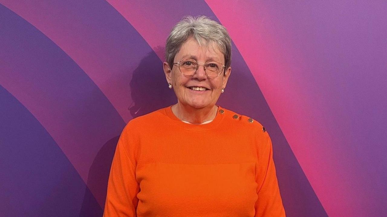 Janet McConkey, a woman wearing glasses and an orange jumper is standing against a prurple background.