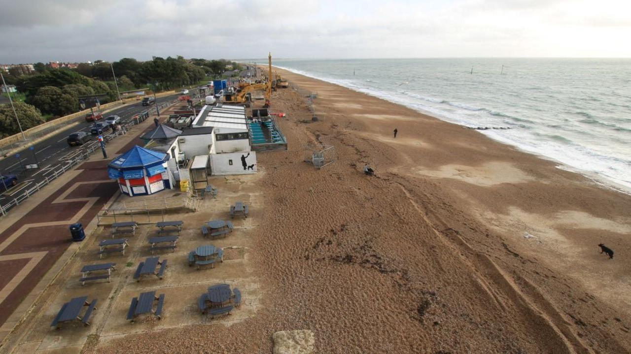 Beach with sea to the right and road and paths to the left.  There are a number of huts and tables and chairs running alongside the rear of the sand.