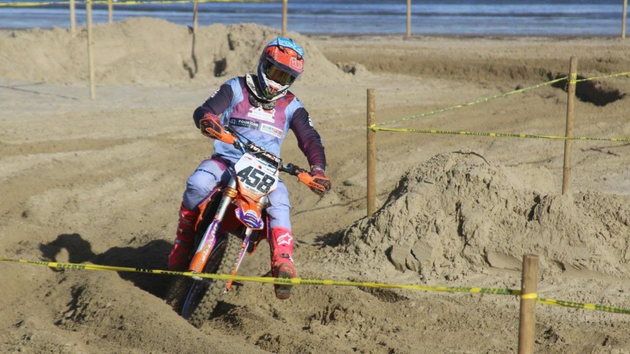 A motocross rider in multi-coloured clothing negotiating a left-hand bend on the sandy track on Weymouth beach. The course is marked out by wooden poles with yellow tape stretched between them.