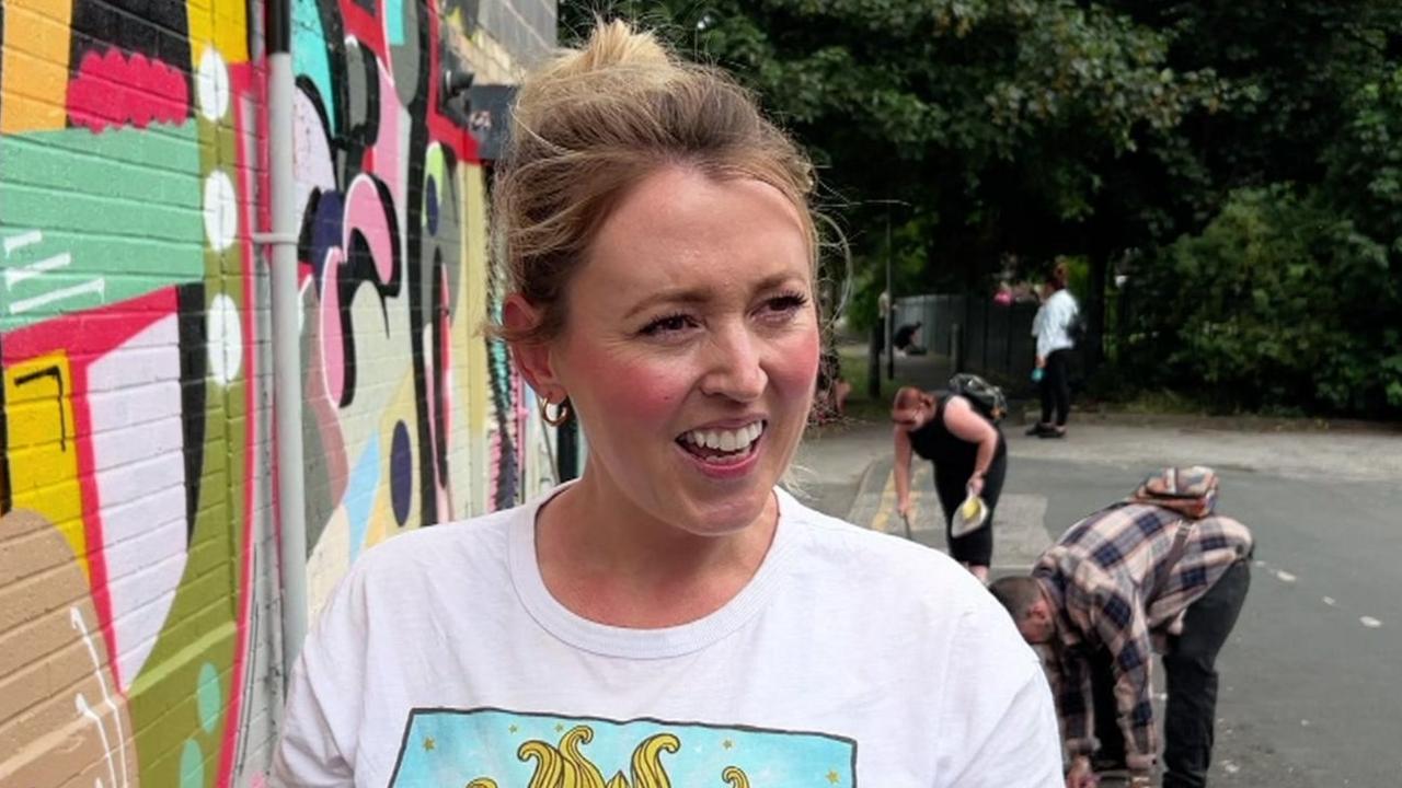 Lucia Thomson wears a white T-shirt and gold hoop earrings, and has her blonde hair tied up. She is standing next to a wall with colour artwork on it and people in the background can be seen sweeping the floor