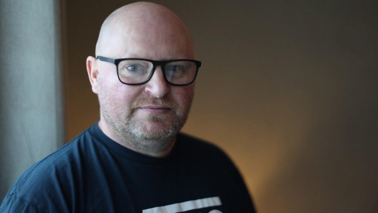 A bald man with dark-rimmed glasses stares at the camera in a dimly lit room. He is wearing a navy t-shirt and has a neutral expression.  