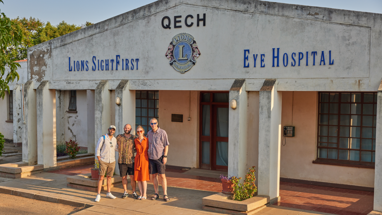 Mrs Patel and her husband kish posing for a photo along two other men in front of a hospital in Malawi. It is a sunny day. The words Lions SightFirst Eye Hospital are displayed at the front of teh building.