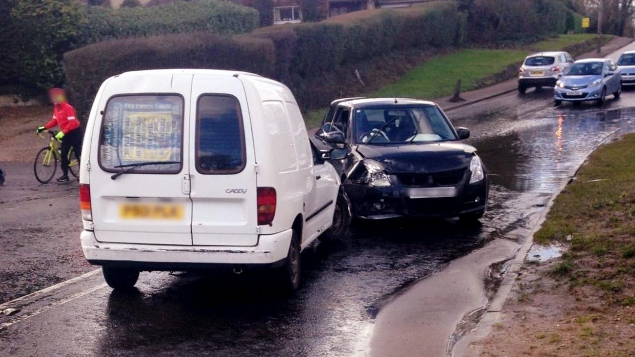 A white van and a black car that have collided on Allum Lane, which has a lot of floodwater on it. There is a cyclist pushing a bicycle in the background, as well as other cars.