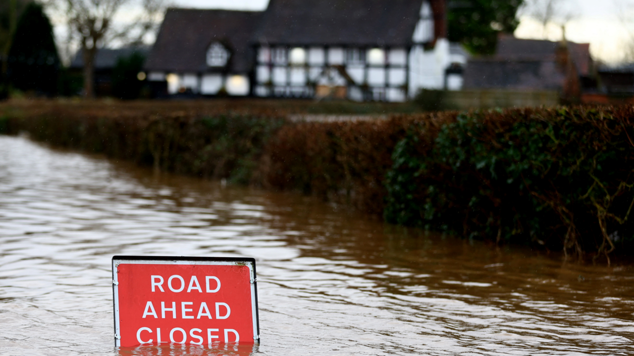 A flood sign in a flooded road saying "road ahead closed". There is a hedge and a black and white house behind the road.