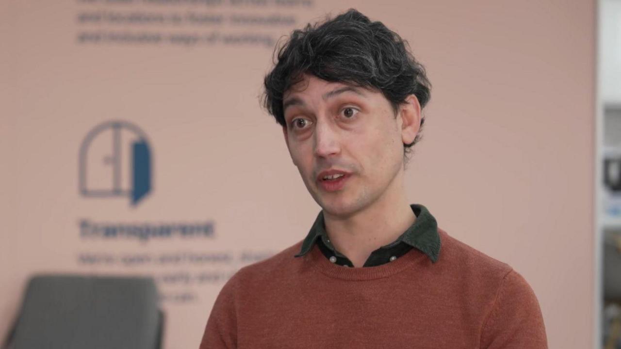 David Mendes da Costa sits speaking to someone off camera in a BBC interview. He has dark hair streaked with grey and is wearing a green shirt and light red jumper.