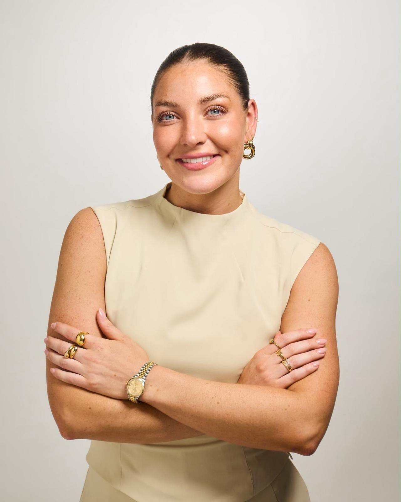 A professional photograph of Daisy Kelly with a slick back bun, wearing a cream sleeveless high neck top with her arms folded and smiling