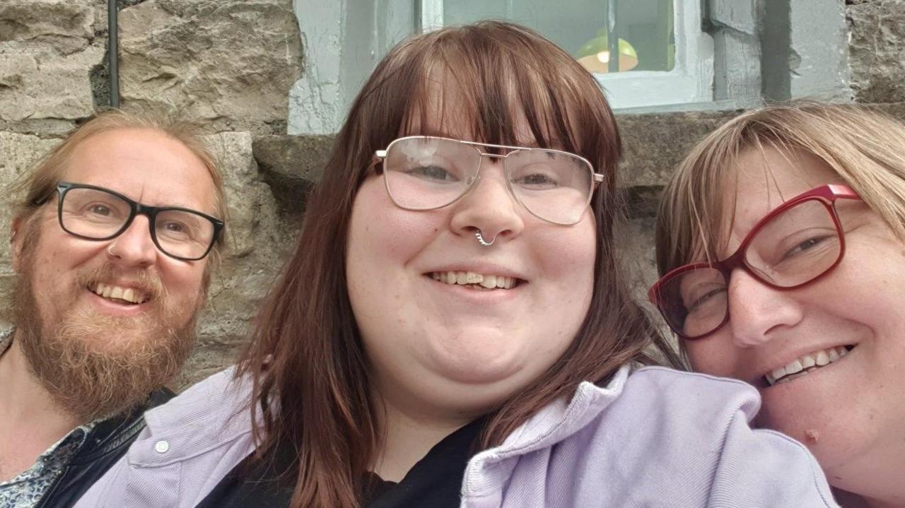 Zoe is pictured between a man and her mum. She has long brown hair, a fringe, glasses, a purple coat, and a nose ring, and is smiling. Mrs Nunn is wearing red glasses and has blonde hair and a fringe. The man also wears glasses and a nose ring, and has ginger hair and a beard.