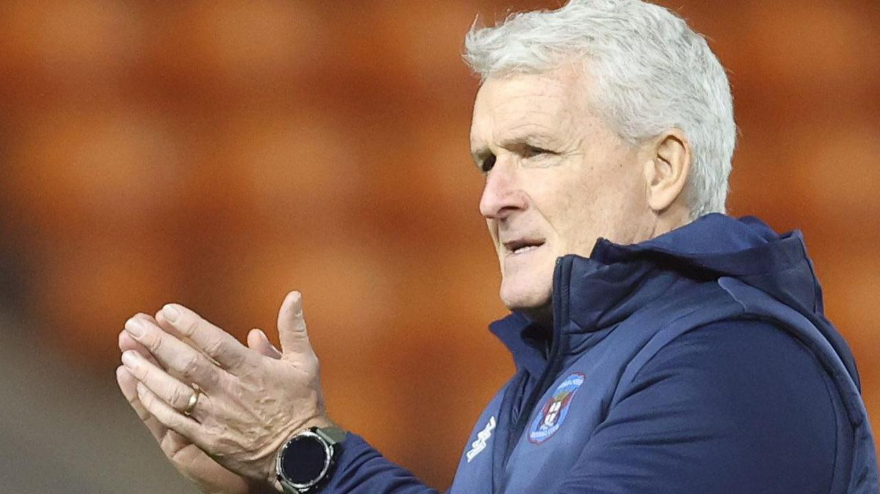 Mark Hughes and another person with a big coat and woolly hat, clap their hands towards Carlisle fans 