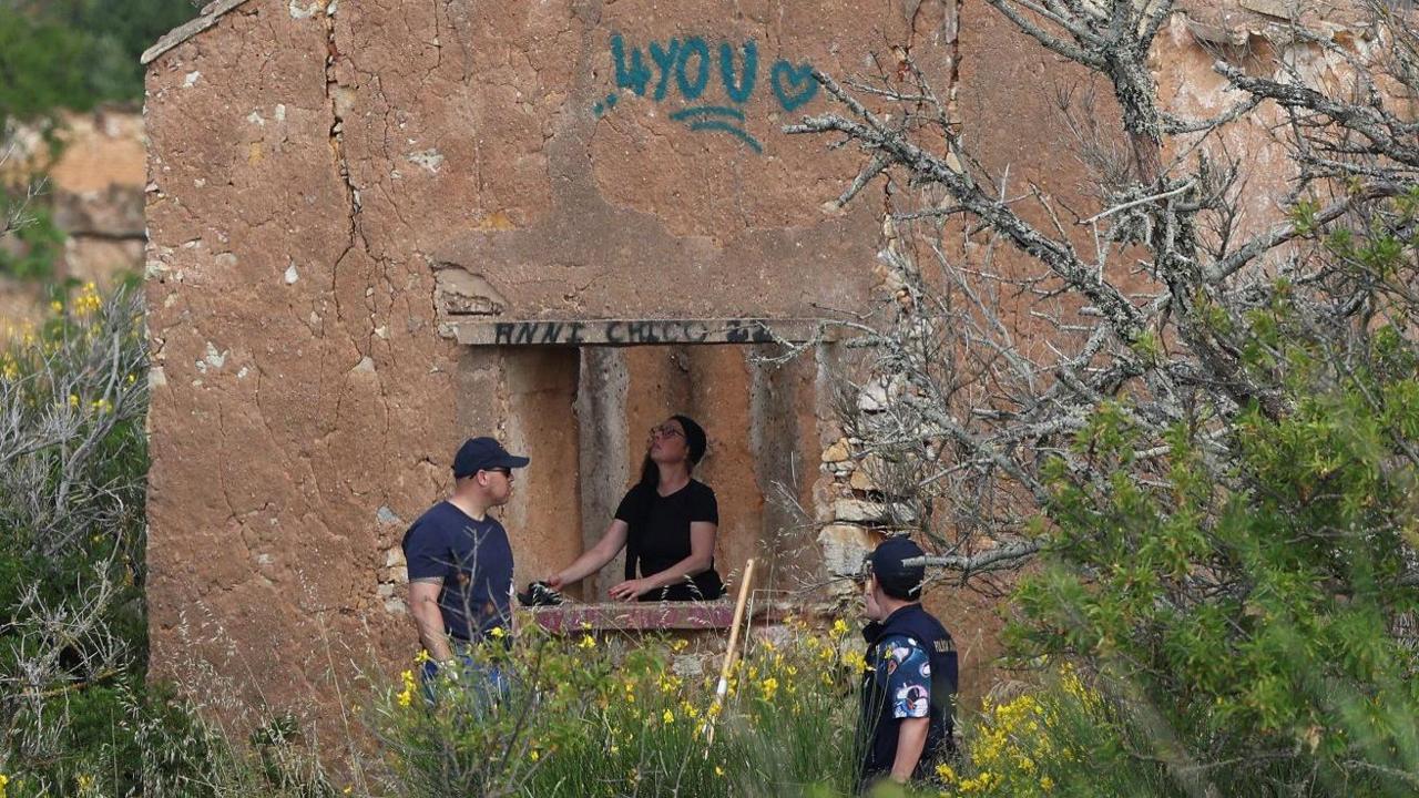 A ruined building. A woman is standing inside the building visible through the hole where a window would have been. Two men are standing outside it. There is green and black graffiti on the building exterior