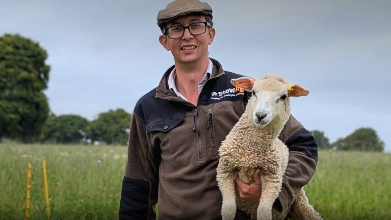 Karl Franklin holds a lamb under his left arm. He wears a brown top, flat cap and glasses. Behind him is a field and trees.