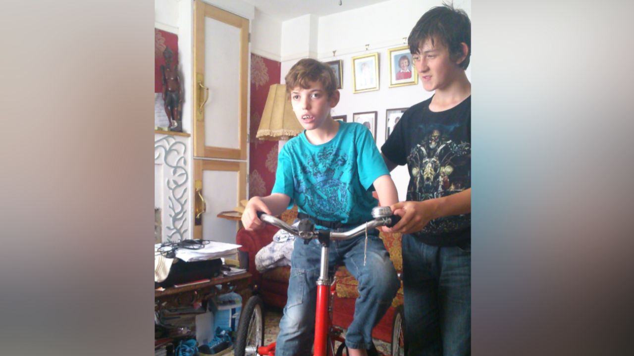 Harry as a child sits on a bike that is held up by his brother. Harry wears a light blue top and jeans. His brother wears a dark T-shirt and jeans.