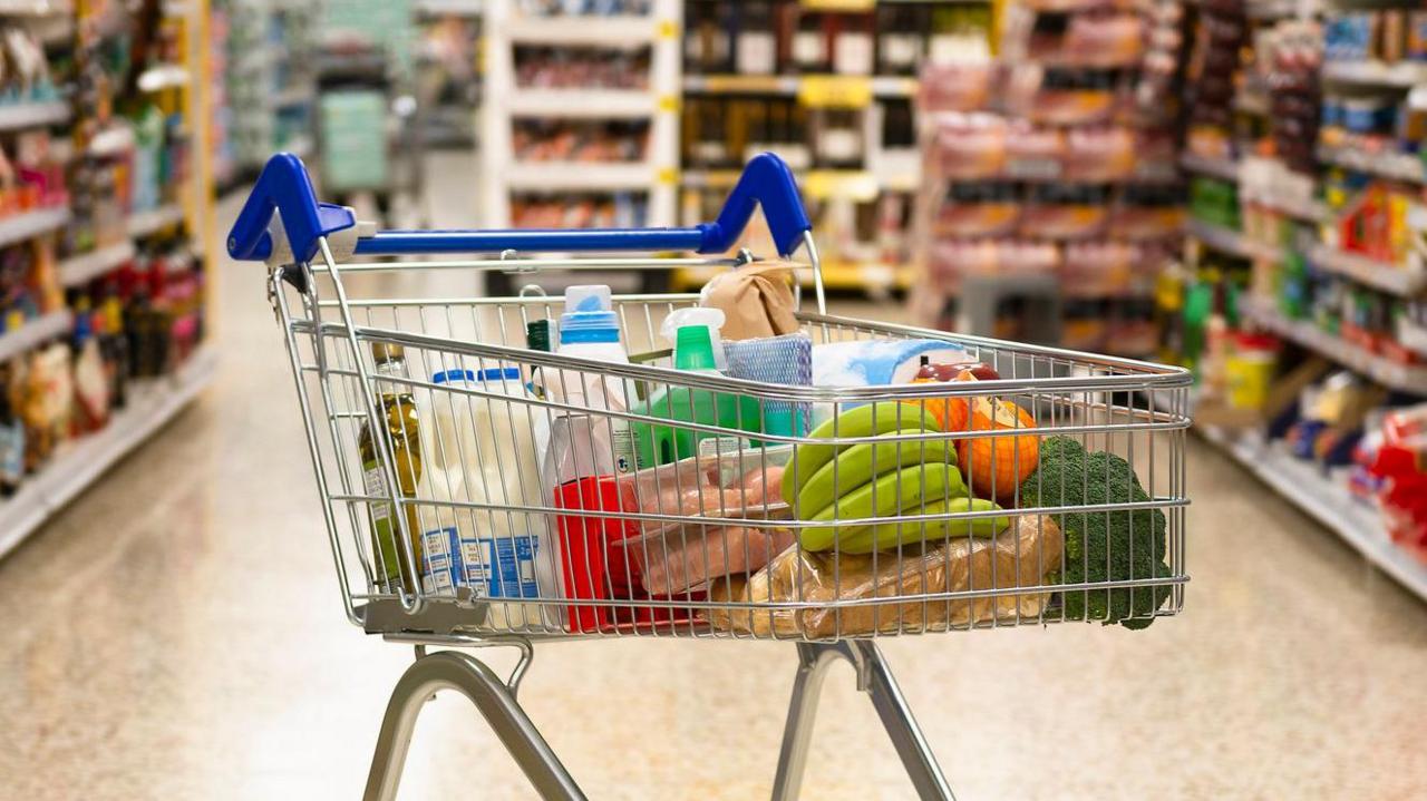 A shopping trolley in the middle of a supermarket aisle. It is filled with grocery items, like milk and bananas. Surrounding the trolley are shelves, but the contents are out of focus.