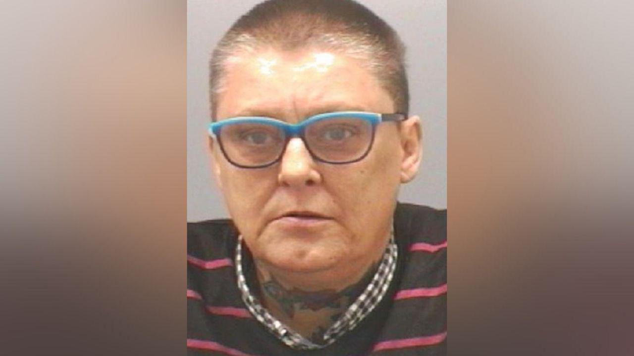 Mugshot of Heslop. She has very short dark shaved hair and blue framed glasses, with a tattoo of flowers across her throat.