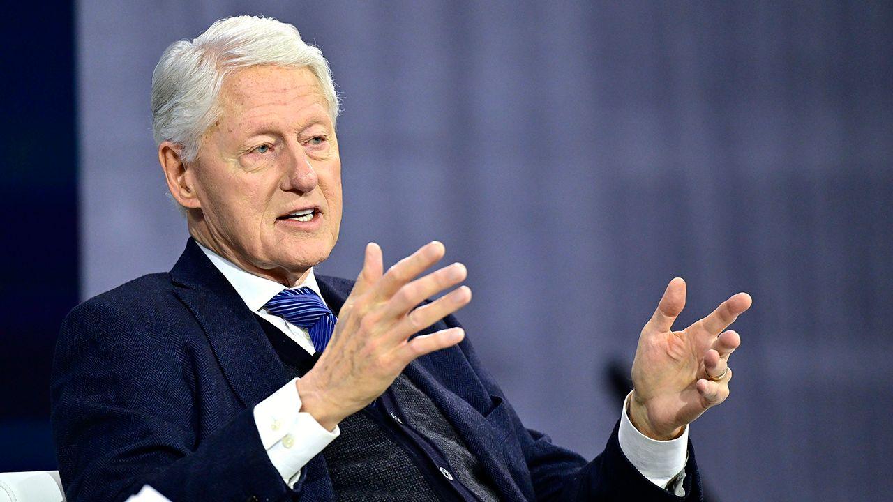 Bill Clinton gesturing while sitting in a chair during a summit in New York on 4 December. He's wearing a suit and tie and looking away from camera. 