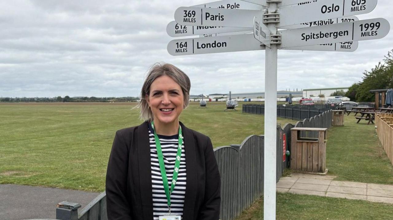 Sophie Parker is standing on an airfield next to a white sign with places across the world written on the sign. She is wearing a black jacket over a white and black striped shirt.