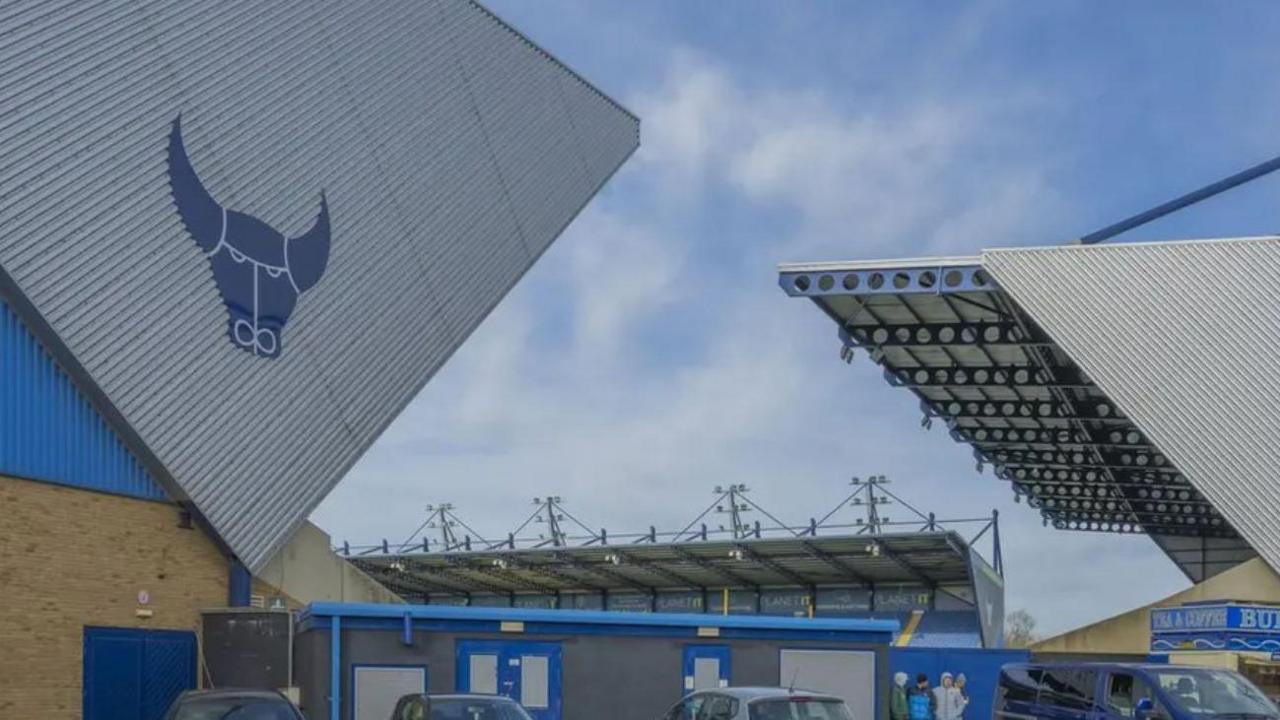 Outside the Kassam Stadium in Oxford on a cloudy day. The signature bull image associated with the team is seen on the side of the stand. There are some people and cars by the building.