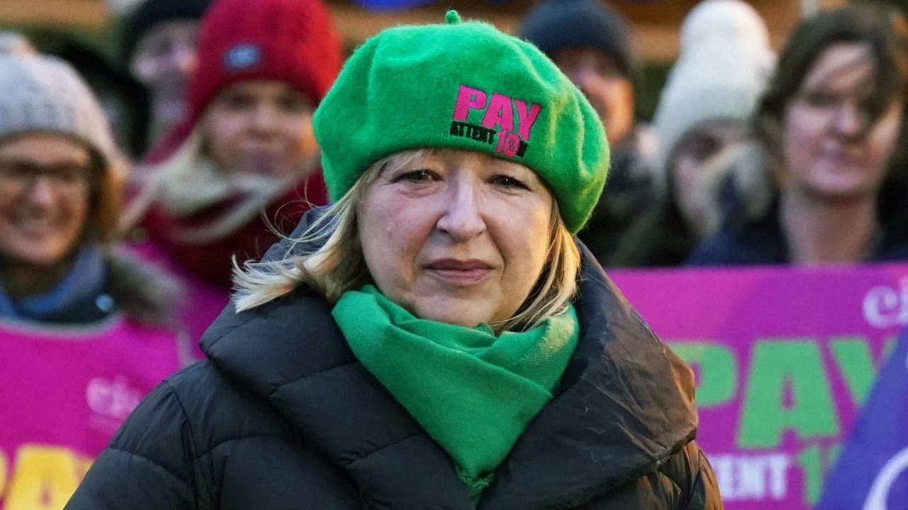A woman with blonde hair on a picket line, wearing a green hat and scarf and a black jacket.