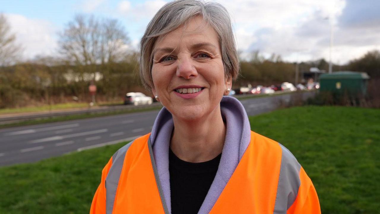 Lilian Greenwood is wearing an orange hi-vis jacket over the top of a coat and black T-shirt. She is standing on a grass verge, with a road in the background.