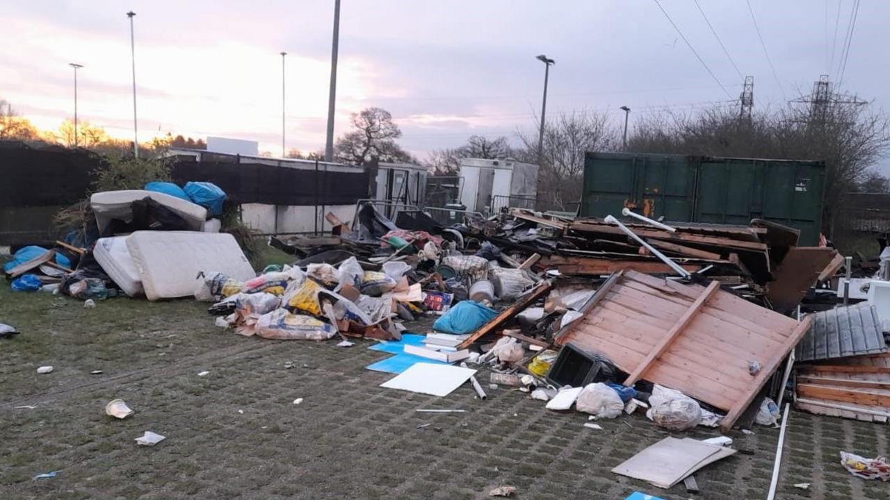 A large amount of rubbish including mattresses, bags of rubbish and wooden hoardings dumped in a car park by a football ground.