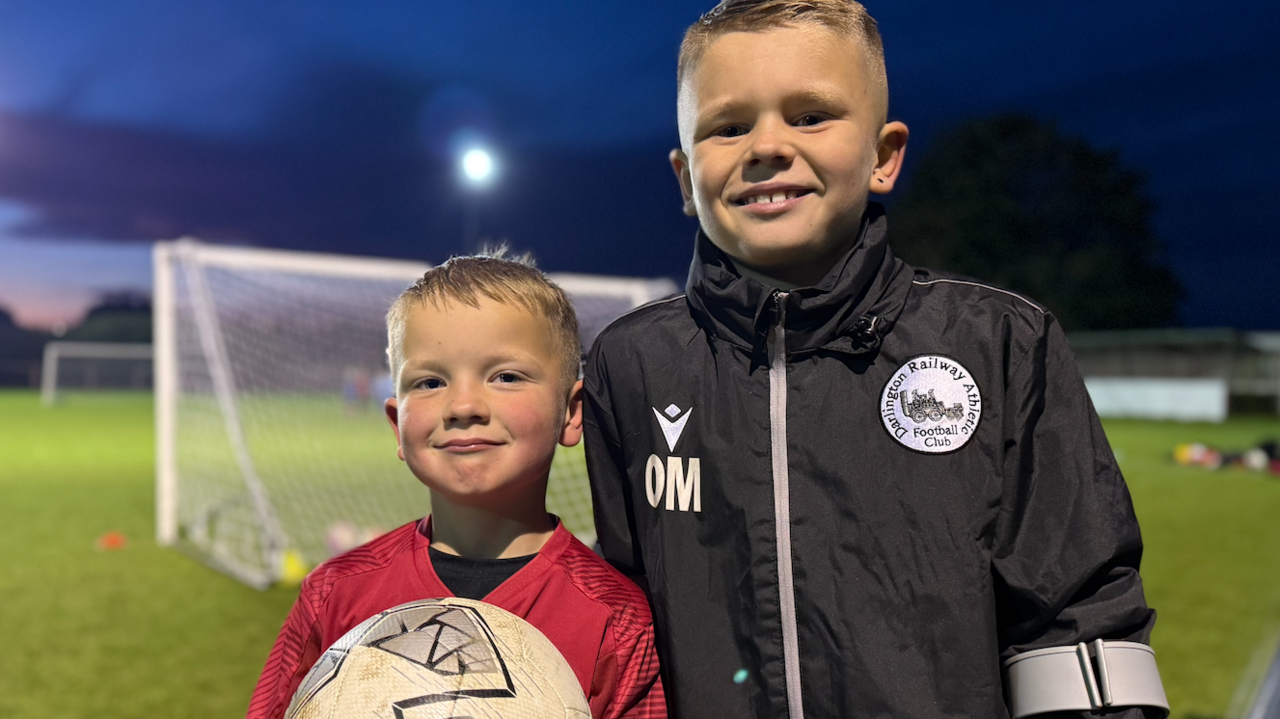 A boy wearing a red shirt holds up a white football while standing next to his brother, who is wearing a black jacket which has a logo saying Darlington Railway Athletic Football Club embossed into it. They are both standing behind a goal post on a grassed football pitch.