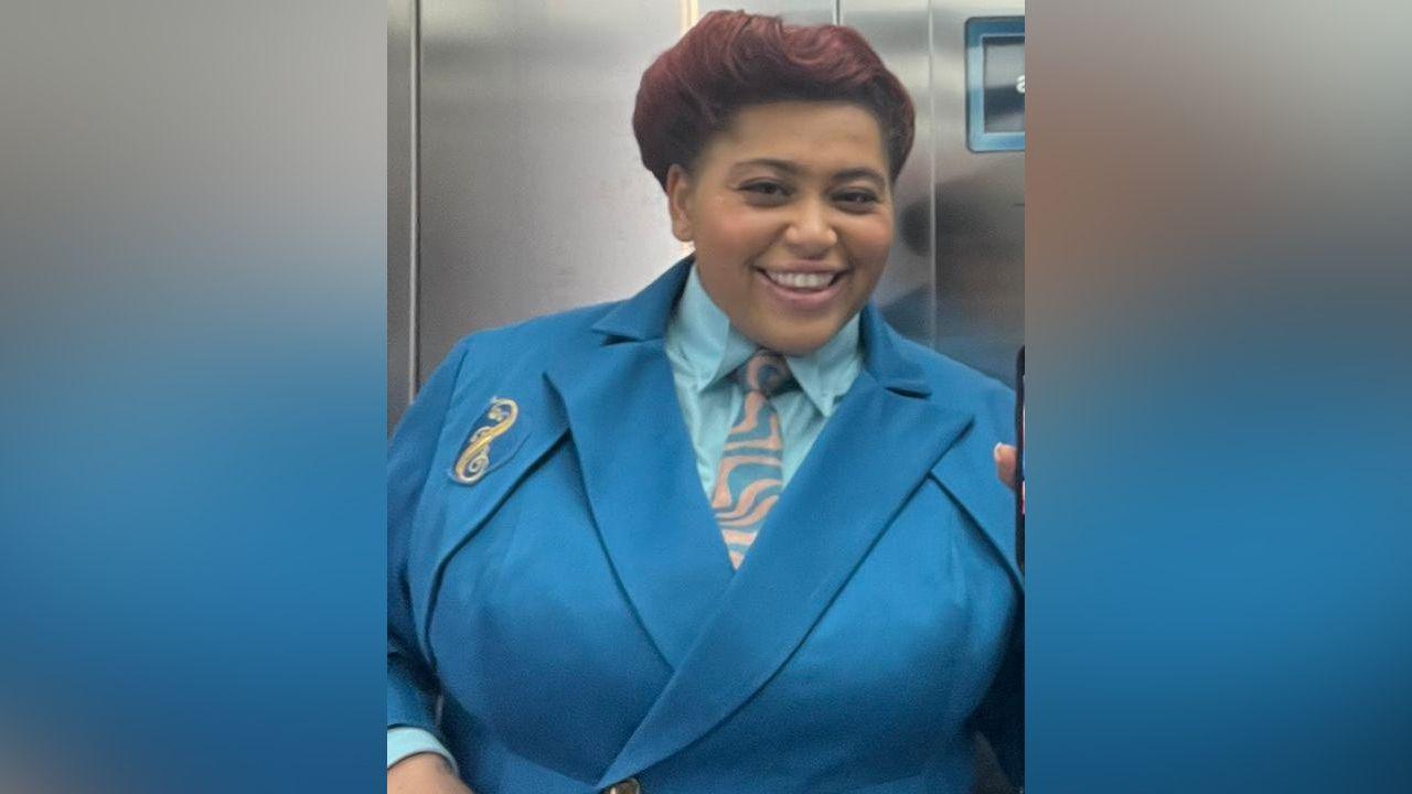 Tegan Browne-Winter is smiling in a mirror selfie she has taken in a lift. She is wearing her Wicked costume which is a bright blue school uniform blazer with a blue shirt underneath. The emblem of Shiz University, a gold S, is on the blazer.