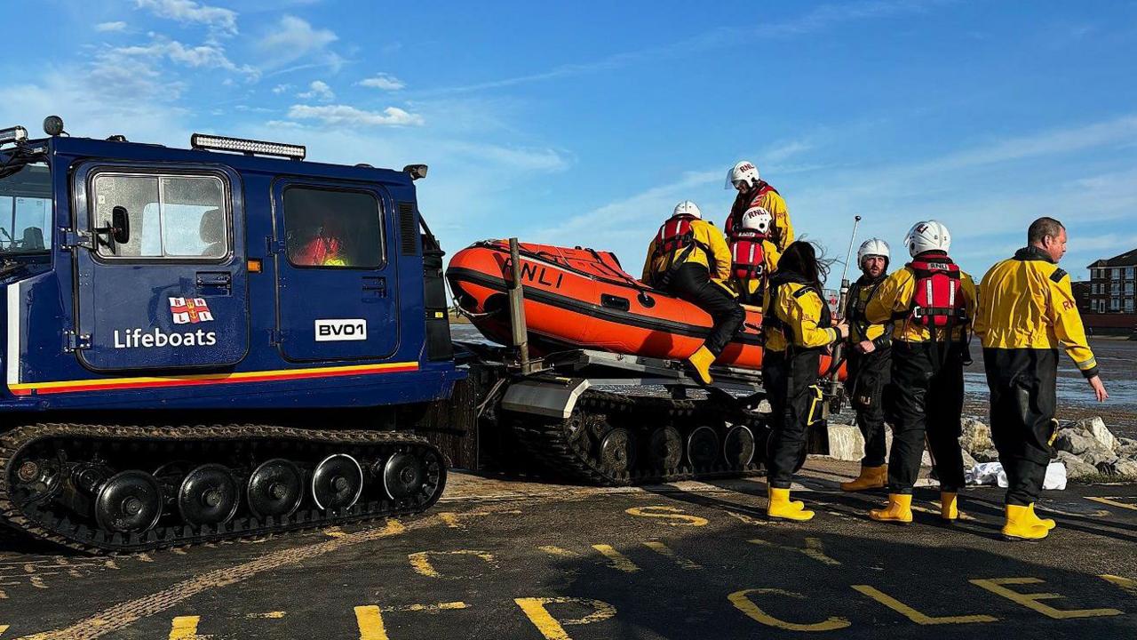 Seven volunteers in yellow jackets and boots surround the lifeboat - which is attached to the back of a blue vehicle - on a dry tarmac area near West Kirby beach