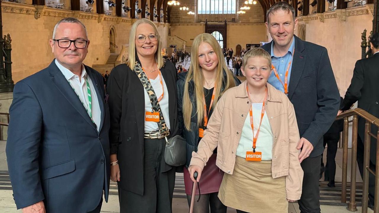Lee Barron, with his eyes closed, standing next to Gemma Blair, Millie's sister, Millie and Gordon Blair. They are in a House of Commons hall, by a staircase. Millie is holding onto a stick. They are all looking at the camera.