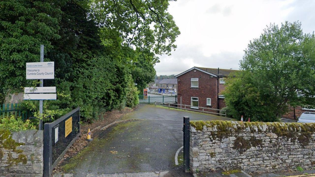 The black entrance gate leading to Cockermouth Coroner's Court. It is a two-storey brown brick building. There is a stone fence and two signs reading Welcome to Cumbria County Council and HM Coroner.