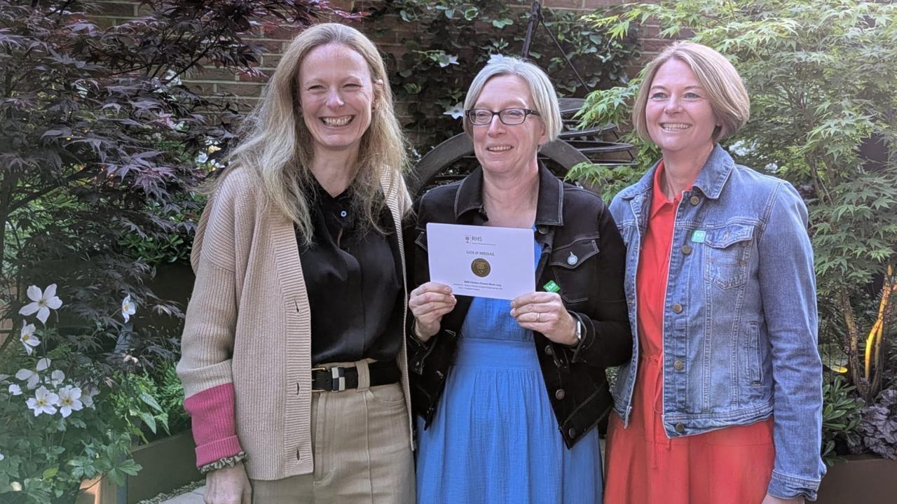 Three women are smiling. A woman in the middle with glasses is holding a paper envelope with the medal encased.