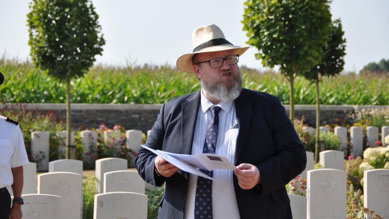 Peter Francis, a bearded man with glasses and wearing a suite and a sunhat, reading from a paper programme. He's standing in front of rows of white graves and it's a sunny day.