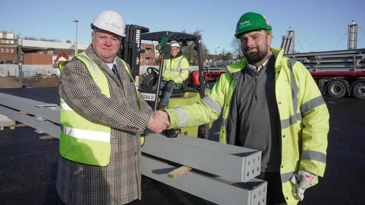 Two men wearing a hard hat and hi-vis jacket are shaking hands and looking at the camera. They are near to two long steel objects.