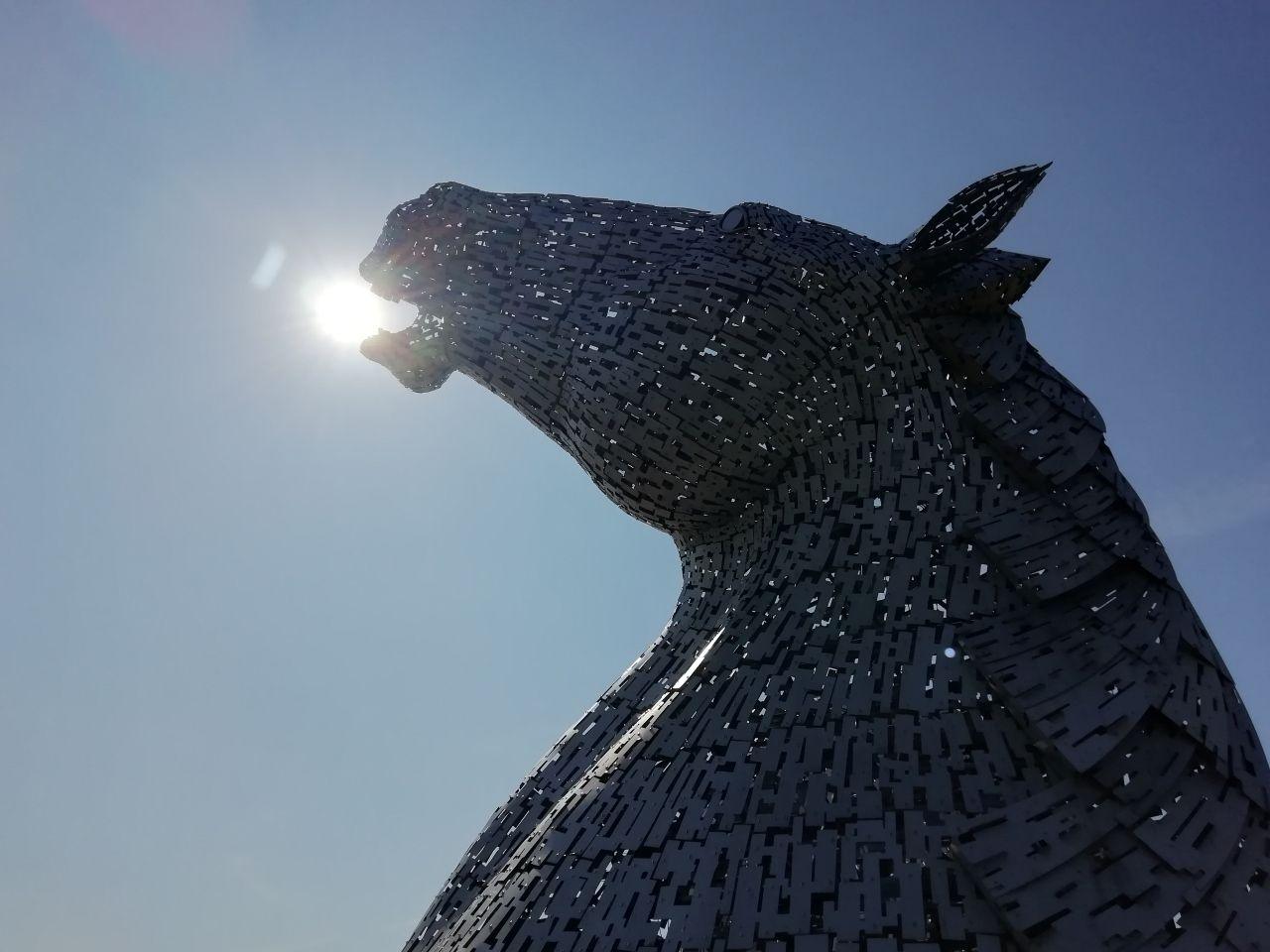 One of the Kelpies, a large metal sculpture of a horse's head, with the sun shining through a gap in its mouth, as if the Kelpie has a ball in its mouth