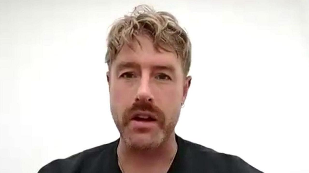 Christian Aldridge talks during a Zoom call. He is sat in front of a white wall. He has short but wavy blonde hair and has a stubbly beard. He is wearing a black t-shirt.