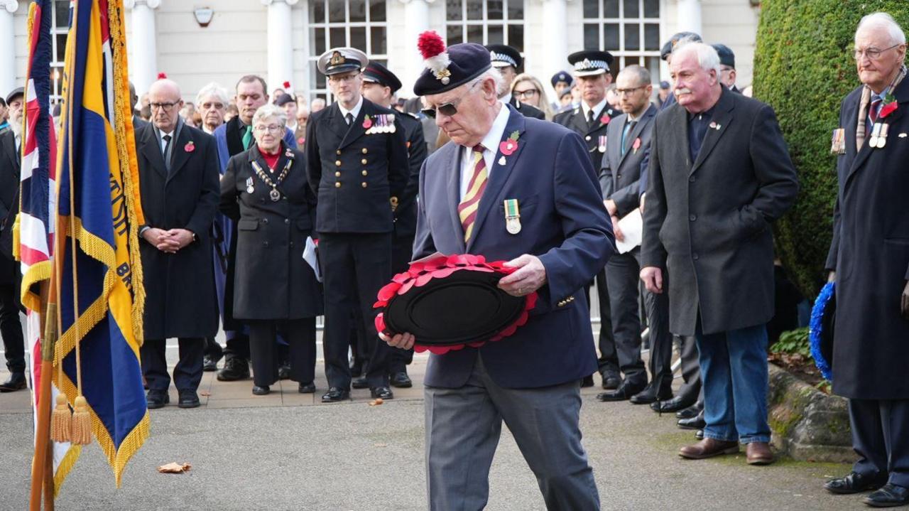 A man with white hair and sunglasses in a blue suit and blue beret, holding a poppy wreath in front of a blue and gold flag, with other people in dark clothing and military uniforms behind him.