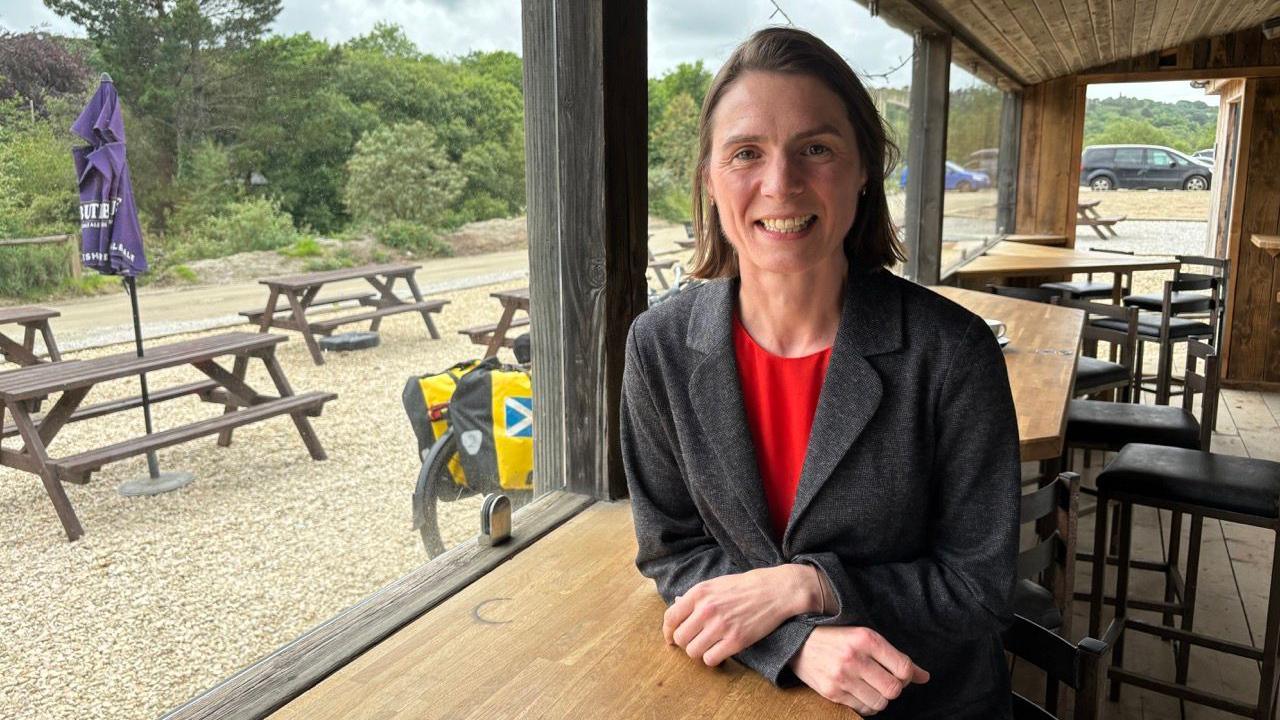 Ruth Gripper sat at a table. She has short brown hair and is wearing a red top and grey coat. Behind her are tables and stool chairs. To the left of her is a window with a pub garden, it has benches and umbrellas. 