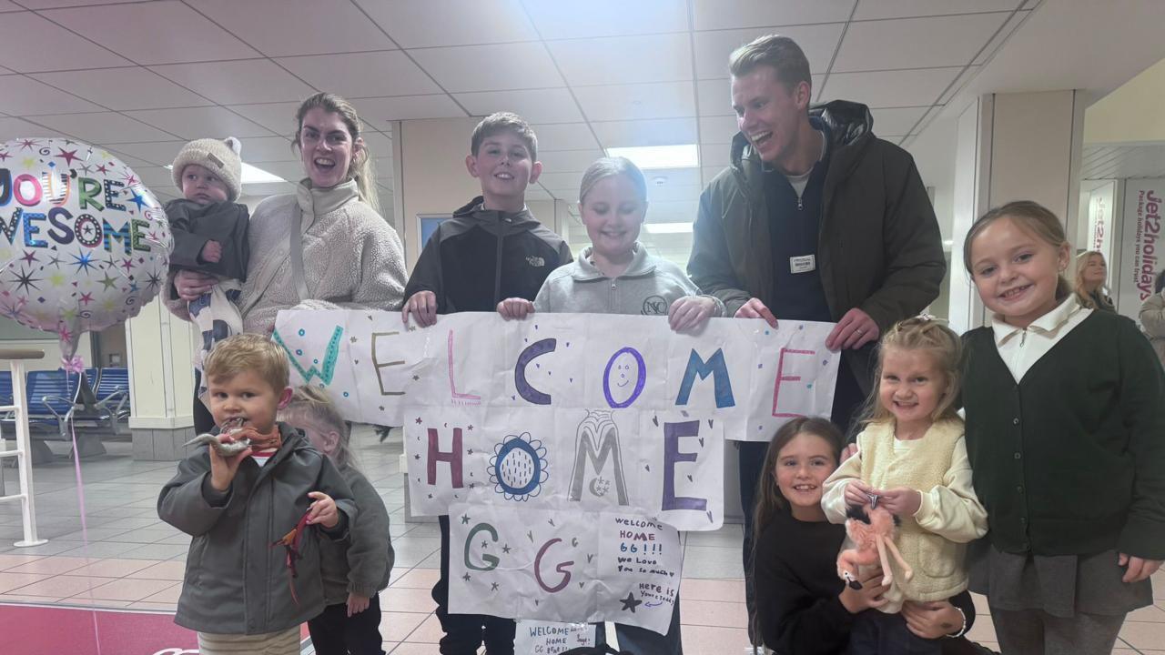 Tom and his family are in Jersey Airport with a paper sign saying "Welcome home GG". There's a balloon which says "You're awesome".