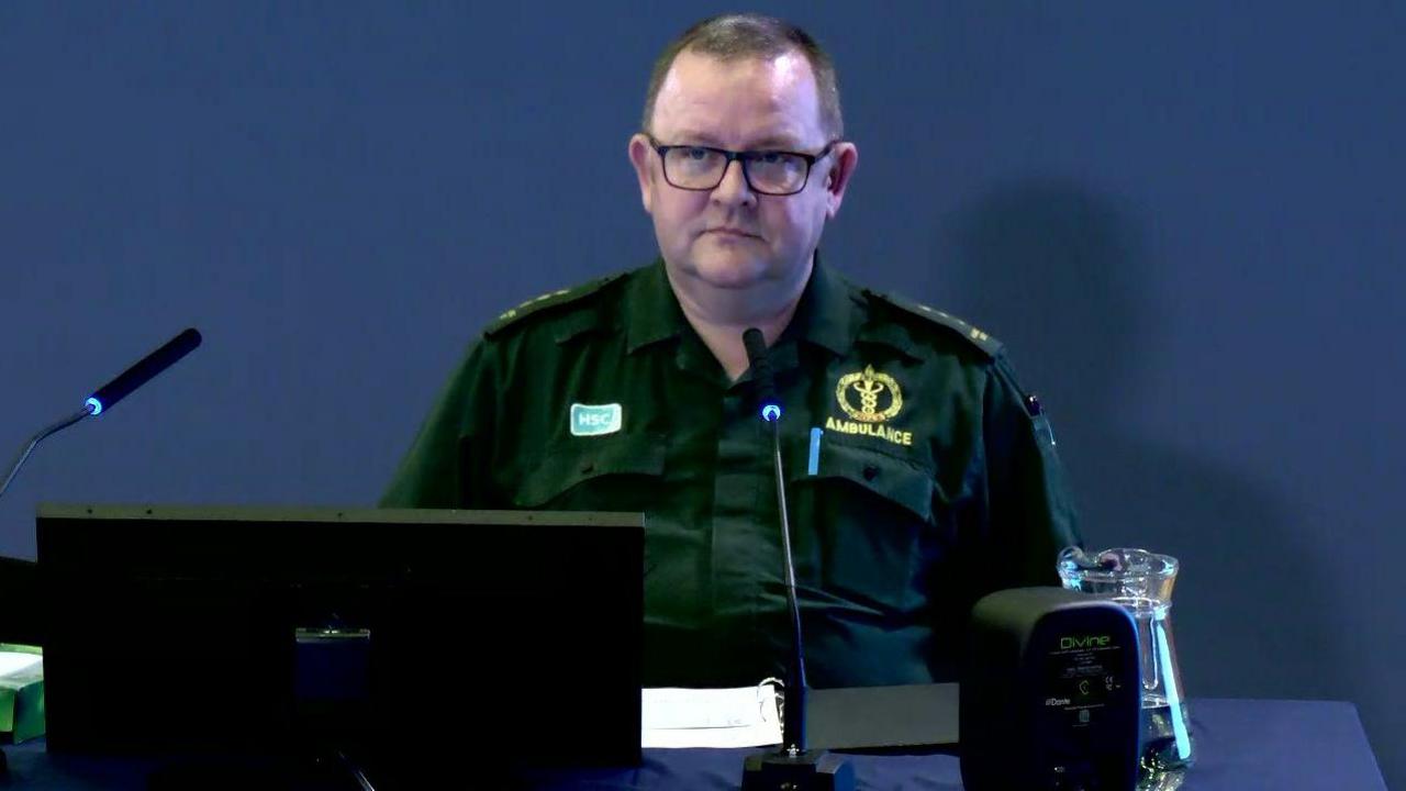 A man is sitting with a computer in front of him. He's wearing a paramedic uniform that says ambulance on it. It's a bottled green colour. A jug of water is sitting on the table in front of him.