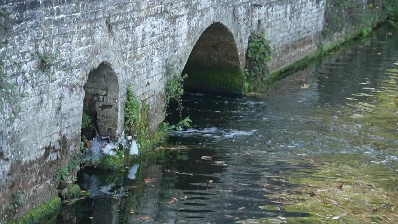 A grey stone bricked bridge was going over a small river. On the right was green algae and on the left, near the bridge, was white foam.