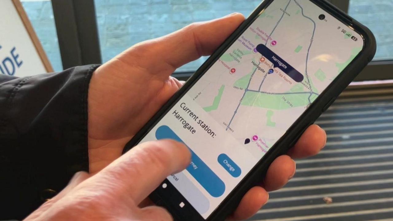 A phone in someone's hand with a screen showing the new system. It has a map with a pin on Harrogate Railway Station and underneath, it says "Current Station: Harrogate".