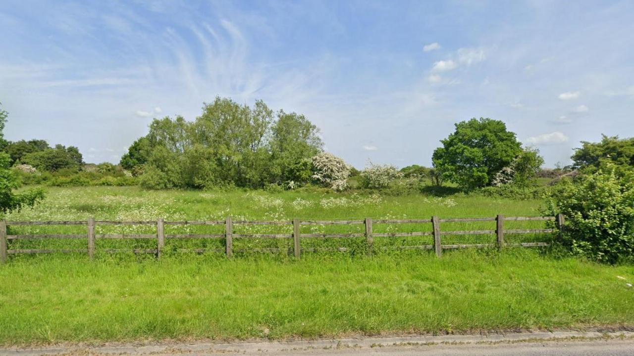 A large patch of land beside the A363. The picture is taken on a sunny day in summer, and the trees and grass is bright green and overgrown. There is a wooden fence blocking off the site from the road.