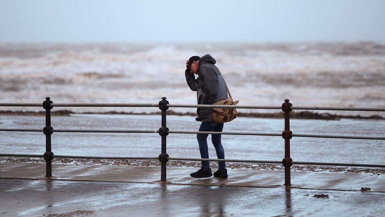 A man clutches his hat while walking along a windy seafront with rough waves in the background