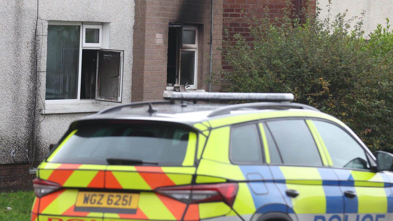 A police care sits outside the door of the arson attack house. The windows are open and the house is scorched.