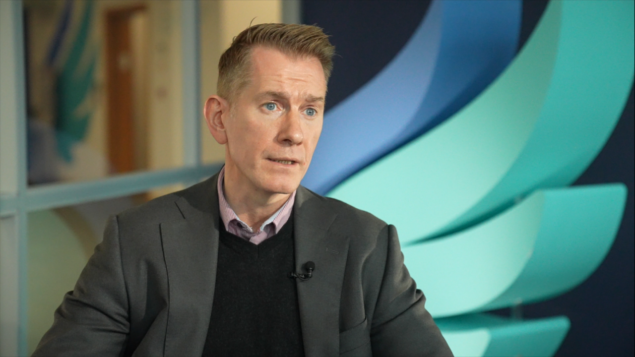 A man in a grey suit with a black jumper, sits in front of a blue winged logo.