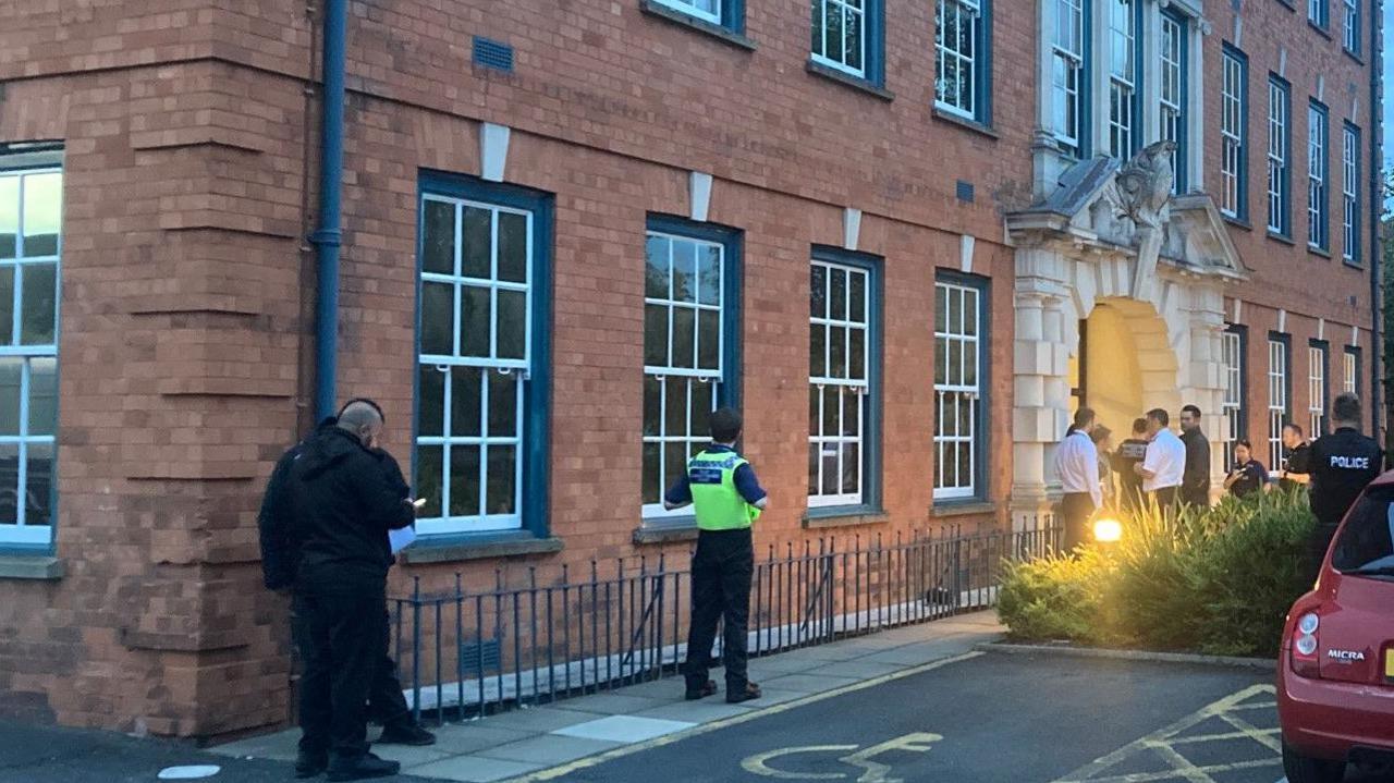 At least half a dozen people are standing outside an entrance to the building, which is on the right of the photo. Three other people are in the middle and left of the image.