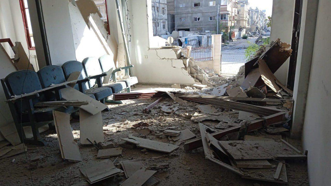 A badly damaged wall of an office, debris is scattered inside it and dust covers a bank of witting room chairs