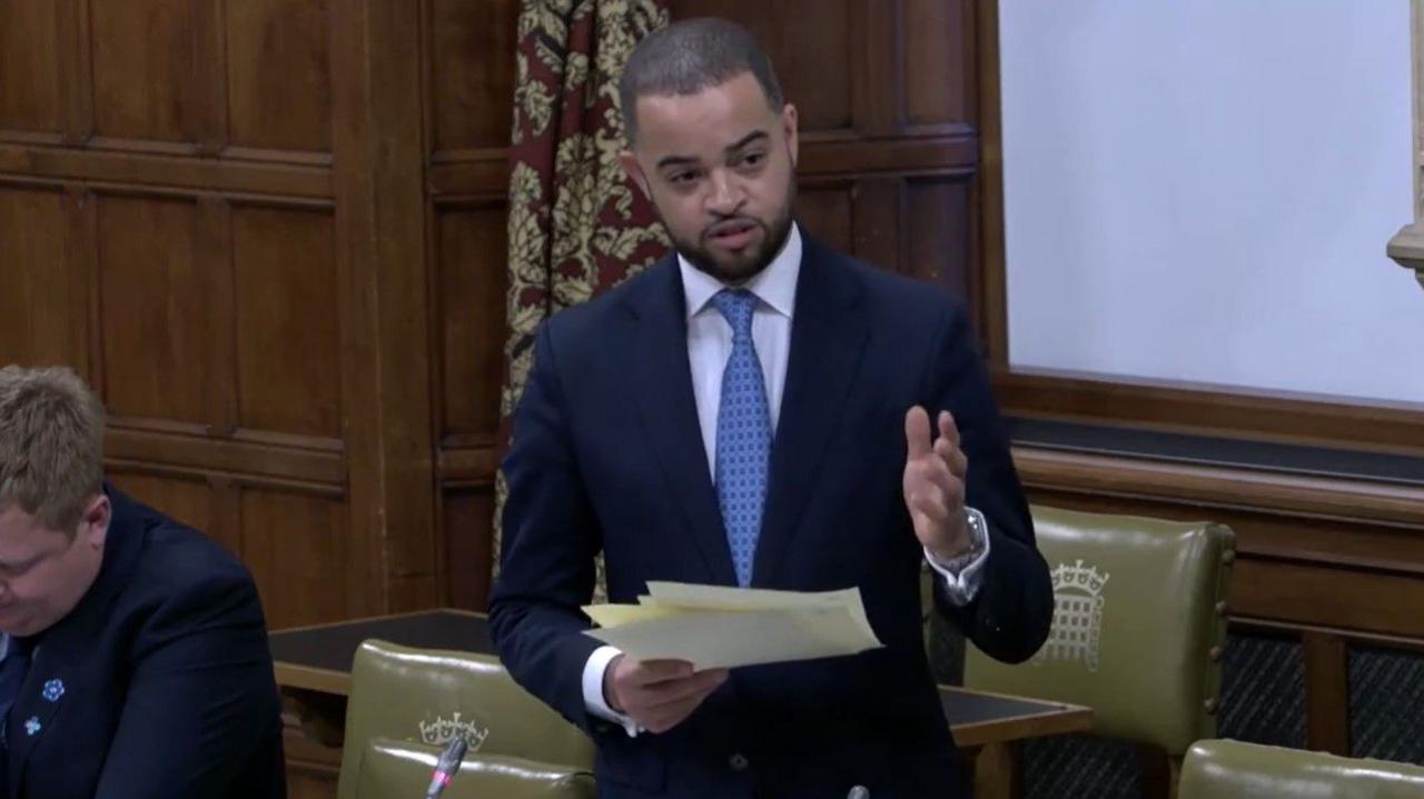 Newcastle-under-Lyme MP Adam Jogee wearing a dark jacket, white shirt and light blue kit. He is standing up during a Westminster Hall debate. 