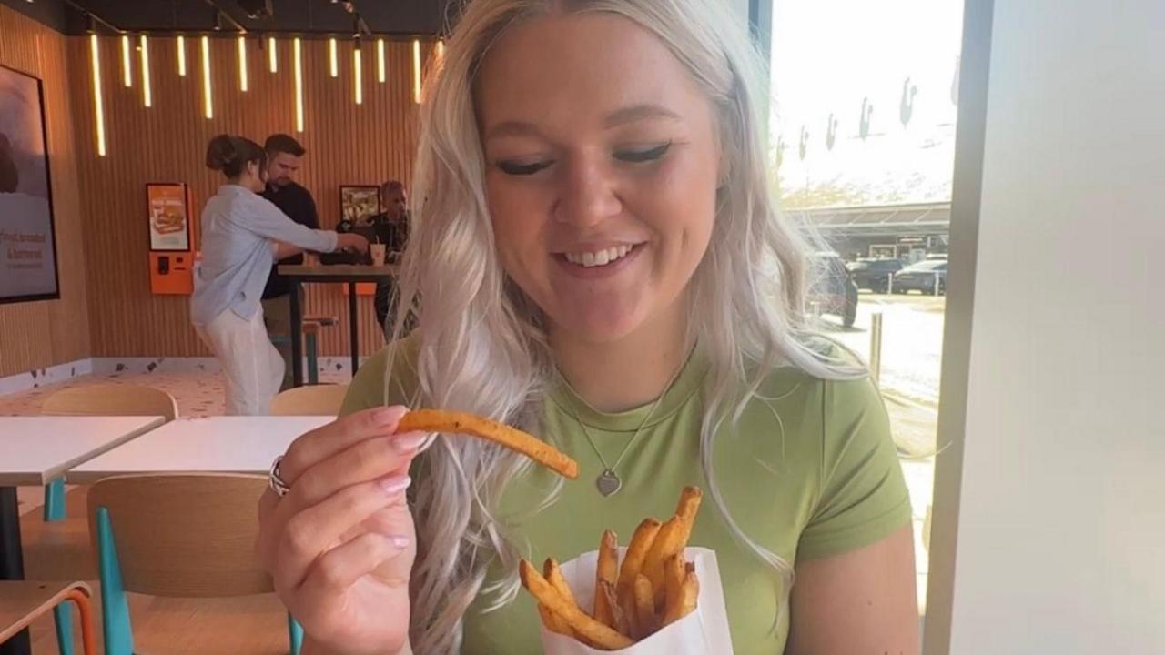 A woman sitting at a table in a resturant eating from a bag of chips. She is looking down at the chips and smiling. She is wearing a green t-shirt.